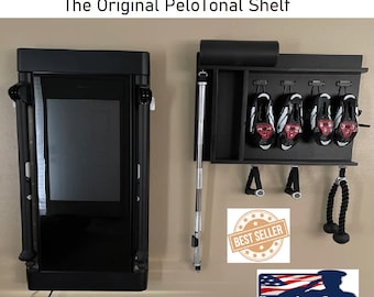 May include: A black wall-mounted shelf with a black Peloton bike stand, a black yoga mat, and a black barbell. The shelf has hooks for hanging items, and there are four pairs of cycling shoes hanging on the hooks. The shelf also has a black rope hanging from it. The text "BEST SELLER" is on a sticker on the shelf. The text "Veteran Owned Business" is on a sticker below the shelf.