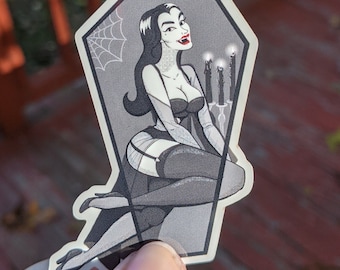 May include: A sticker featuring a woman in a coffin with a spiderweb and candles. She is wearing a black dress and has long black hair. The sticker is on a white background.