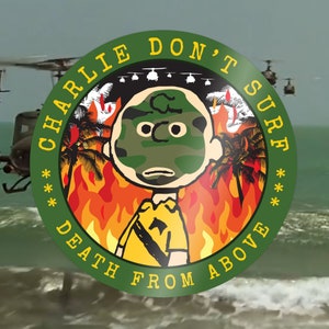 May include: A circular green graphic with the text "CHARLIE DON'T SURF DEATH FROM ABOVE" surrounds a cartoon character with a camouflage face. The background shows helicopters, flames, and palm trees.