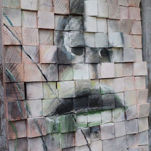 Puede incluir: Un primer plano de una pieza de arte de pared de madera con un mosaico de bloques de madera pintados. Los bloques están dispuestos en un patrón de cuadrícula y pintados en tonos de gris, negro, blanco y verde. La obra de arte representa una cara con bigote.