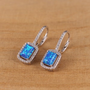Puede incluir: Un par de pendientes de plata con piedras de ópalo azul engastadas en un marco cuadrado con un halo de piedras más pequeñas. Los pendientes tienen un cierre de palanca.