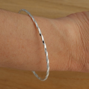 Könnte beinhalten: Ein silbernes, gedrehtes Armreif-Armband. Das Armband besteht aus einem einzigen Stück Metall, das zu einer Spiralform gedreht ist.