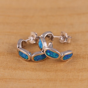 Puede incluir: Un par de pendientes de aro plateados con incrustaciones de ópalo azul de forma ovalada. Los pendientes se exhiben sobre una superficie de madera, mostrando su metal pulido y los vibrantes detalles de las piedras preciosas. Son una joya.