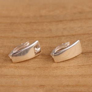 Puede incluir: Un par de pendientes de aro rectangulares plateados. Los pendientes tienen una superficie pulida y reflectante y un diseño simple y moderno. Se muestran sobre una superficie de madera.