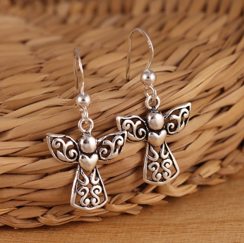 May include: A pair of silver dangle earrings featuring an angel design with intricate details. The angel has a heart-shaped body and delicate wings.