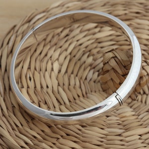 Op de afbeelding: Een zilveren armband met een scharniergesp. De armband ligt op een geweven rieten oppervlak.
