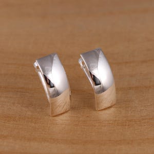 Puede incluir: Un par de pendientes de aro plateados y brillantes. Los pendientes tienen forma rectangular con bordes redondeados y un acabado pulido. Los pendientes se muestran sobre una superficie de madera de color marrón claro.