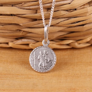 May include: A silver Saint Christopher pendant necklace. The round pendant features an engraved image of Saint Christopher carrying a child, with the words "SAINT CHRISTOPHER" around the edge. The necklace has a delicate chain.