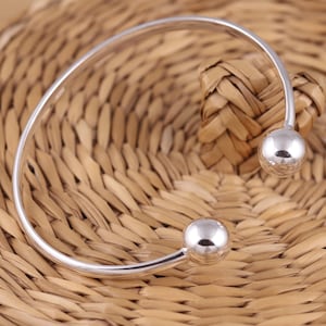 May include: A silver open cuff bracelet with a smooth, rounded design. The bracelet has a spherical ball at each end. The jewellery is displayed on a woven, light brown surface.