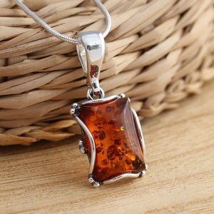 Op de afbeelding: Een zilveren ketting met een rechthoekige hanger van barnsteen. De steen is diep oranje van kleur met bruine en zwarte insluitsels. De hanger is gezet in een zilveren frame met een decoratief ontwerp.