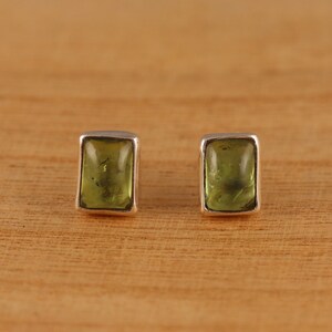 May include: A pair of square-shaped stud earrings. Each earring features a light green, rectangular gemstone set in a silver-colored metal frame. The earrings are displayed on a wooden surface.