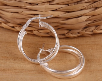 May include: Silver hoop earrings with a twisted design. The earrings are made of a shiny metal and have a classic, elegant look. The earrings are shown on a light brown wooden surface with a woven basket in the background.