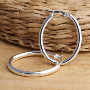May include: A pair of silver oval hoop earrings. The earrings are made of sterling silver and have a polished finish. The earrings are marked with the number 925.