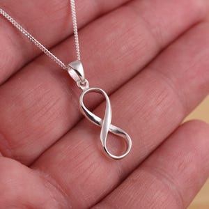 Può includere: Un ciondolo a forma di simbolo dell'infinito argentato su una catena sottile. Il ciondolo è liscio e lucido, riflettendo la luce. La collana è presentata su uno sfondo neutro. La catena è di design delicato.