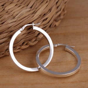 May include: A pair of silver hoop earrings with a flat, wide design. The earrings are simple and elegant.