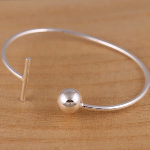 May include: A silver-colored open cuff bracelet with a minimalist design. The bracelet features a small, round ball on one end and a straight bar on the other. The jewelry is set against a light brown wooden surface.
