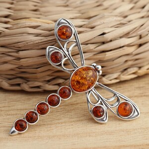 May include: A silver-toned dragonfly brooch with amber-colored stones. The dragonfly has an oval amber stone at its center, with smaller round stones along the body and at the wing tips. The brooch is set against a wooden surface.