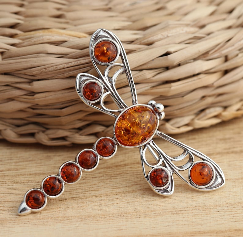 May include: A silver-toned dragonfly brooch with amber-colored stones. The dragonfly has an oval amber stone at its center, with smaller round stones along the body and at the wing tips. The brooch is set against a wooden surface.