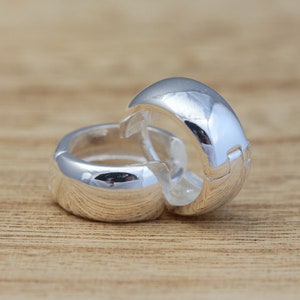 Puede incluir: Dos pendientes de aro plateados con una superficie pulida y reflectante. Los pendientes están colocados sobre una superficie de madera marrón claro, mostrando su diseño geométrico redondeado. Los pendientes están diseñados para ser usados en la oreja.