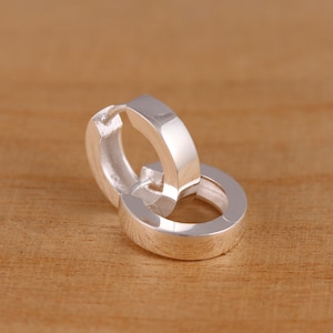 Peut inclure: Deux boucles d'oreilles créoles en argent. Les boucles d'oreilles sont en métal brillant et réfléchissant. Les créoles ont un design simple et classique, avec une forme plate et rectangulaire. Les boucles d'oreilles sont présentées sur une surface en bois.
