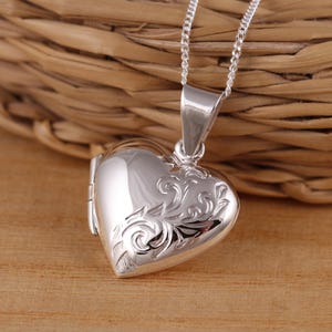 Puede incluir: Un relicario de plata en forma de corazón con un intrincado diseño floral. El relicario tiene una abertura con bisagras y una cadena.