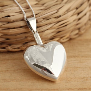 Può includere: Una collana con un ciondolo a forma di cuore in argento. Il ciondolo è vuoto e ha un coperchio a vite. La collana è su una catena d'argento.