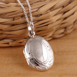 May include: A silver oval locket necklace with a delicate chain. The locket has a detailed engraved design around the edge.