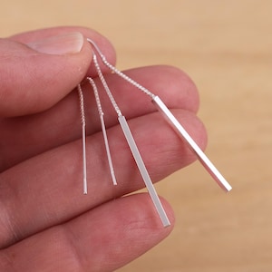 May include: A set of silver bar earrings. The earrings are held in a hand and feature a chain and a straight bar design. The bars are of varying lengths, with a polished finish. The earrings are a simple, modern style.