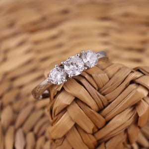 以下が含まれることがあります： 3つの丸い透明な宝石が付いたシルバーリング。リングは、織られた薄茶色の表面に置かれています。宝石が焦点となり、光を捉え、輝きを生み出しています。リングのバンドは細く、磨かれています。