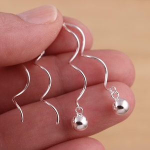 May include: A pair of silver-colored, spiral drop earrings. Each earring features a curved, wavy design with a small, round ball at the end. The earrings are held in a hand, showcasing their delicate and elegant appearance.