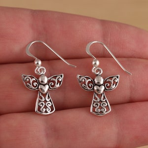 May include: A pair of silver angel dangle earrings with a heart detail. The earrings have a delicate, intricate design.