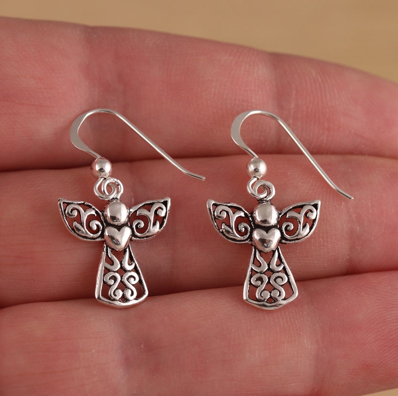 May include: A pair of silver angel dangle earrings with a heart detail. The earrings have a delicate, intricate design.