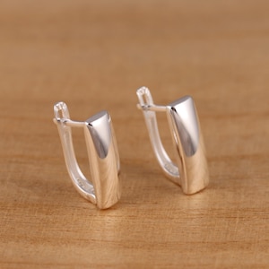 Może przedstawiać: Para błyszczących, srebrnych kolczyków. Każdy kolczyk ma prostokątny, lekko zakrzywiony kształt z zawiasowym zapięciem. Kolczyki są prezentowane na jasnobrązowej drewnianej powierzchni.