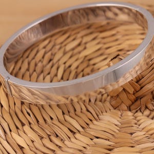 Op de afbeelding: Een zilverkleurige armband met een plat, rechthoekig ontwerp. De armband is gemaakt van metaal en heeft een scharnier. De armband ligt op een geweven, lichtbruin oppervlak.