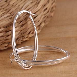 May include: A pair of polished silver oval hoop earrings. The earrings have a smooth, reflective surface and a classic design. The hoops are displayed against a natural wood grain surface and a woven background.