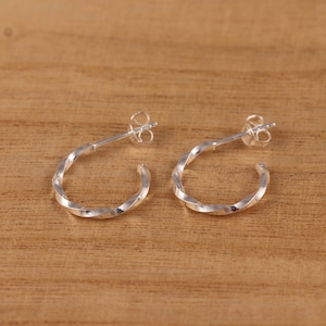 Puede incluir: Un par de pendientes de aro retorcidos de plata. Los pendientes son pequeños y delicados, con un diseño sencillo.
