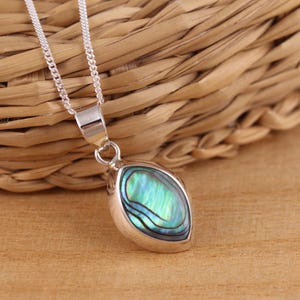 Op de afbeelding: Een zilveren ketting met een ovale abalone schelpen hanger. De schelp toont iriserende blauwe, groene en paarse kleuren. De hanger is gezet in een gepolijste zilveren lijst en hangt aan een fijne zilveren ketting.