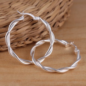 Puede incluir: Un par de pendientes de aro de plata con un diseño trenzado. Los pendientes están hechos de un alambre grueso y trenzado y tienen un aspecto simple y elegante.