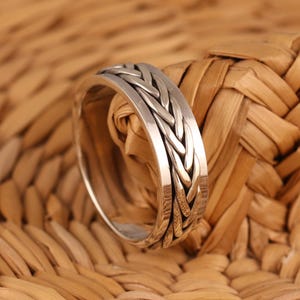May include: A silver ring with a braided design. The ring has a polished finish and a textured, woven pattern around the band. The ring is set against a woven, light brown background.