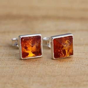 May include: A pair of square amber stud earrings with a silver setting. The amber has a warm, golden-brown color with visible inclusions. The earrings are displayed on a light brown wooden surface.