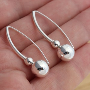 May include: A pair of silver earrings. Each earring has a large, reflective sphere hanging from a curved, elongated bar. A smaller sphere sits above the larger one. The earrings are designed for pierced ears.