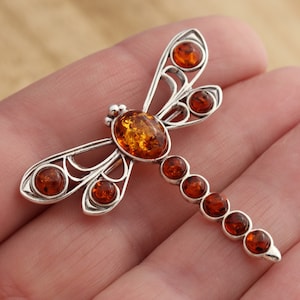 May include: Silver-toned dragonfly brooch with amber-colored stones. The brooch features a detailed design with a large amber stone at the center of the body and smaller stones along the wings and tail. The wings have an openwork design.