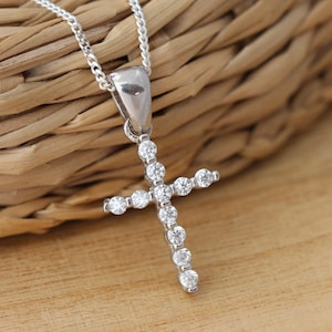 Può includere: Un ciondolo a forma di croce in argento con piccoli diamanti trasparenti incastonati nel metallo. La croce è su una catena d'argento.