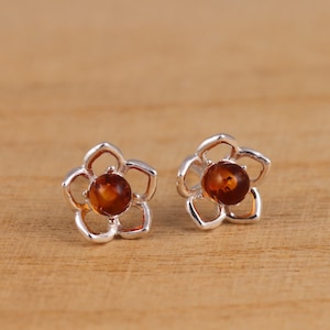 Può includere: Un paio di orecchini a bottone a forma di fiore. Ogni orecchino presenta un design a fiore traforato color argento con cinque petali. Il centro di ogni fiore contiene una pietra rotonda color ambra. Gli orecchini sono esposti su una superficie di legno.