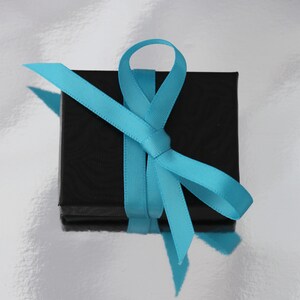May include: A small, square black gift box tied with a bright turquoise ribbon. The ribbon is neatly knotted on top, with the ends of the ribbon hanging down. The box sits on a shiny, silver surface, creating a contrast between the colors.