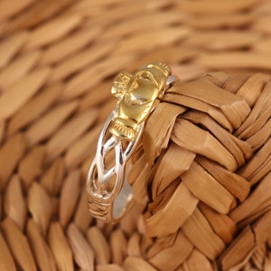 May include: A gold and silver Celtic knot ring featuring a Claddagh design. The ring is displayed against a woven wicker background.