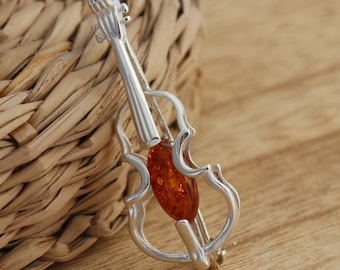 Op de afbeelding: Een zilveren viool broche met een amber steen in het midden. De broche ligt op een geweven rieten oppervlak.