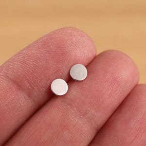 May include: A pair of small, round, silver earrings. The earrings are simple and minimalist in design.