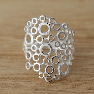 Könnte beinhalten: Ein silberner Ring mit einem einzigartigen offenen Design aus miteinander verbundenen Kreisen unterschiedlicher Größe. Der Ring hat eine polierte Oberfläche und liegt auf einer Holzoberfläche. Das Design ist modern und geometrisch.