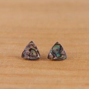 Könnte beinhalten: Ein Paar dreieckige Ohrstecker aus Abalone-Muscheln. Die Ohrringe haben einen silberfarbenen Metallrand und zeigen schillernde Farben von Grün, Blau und Lila. Die Ohrringe sind auf einer hellbraunen Holzoberfläche platziert.